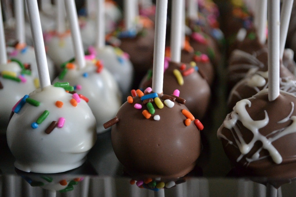 Colorful candy-coated cake pops with sprinkles, white and milk chocolate coating, and decorative white chocolate drizzle, perfect for sweet celebrations and party favors.