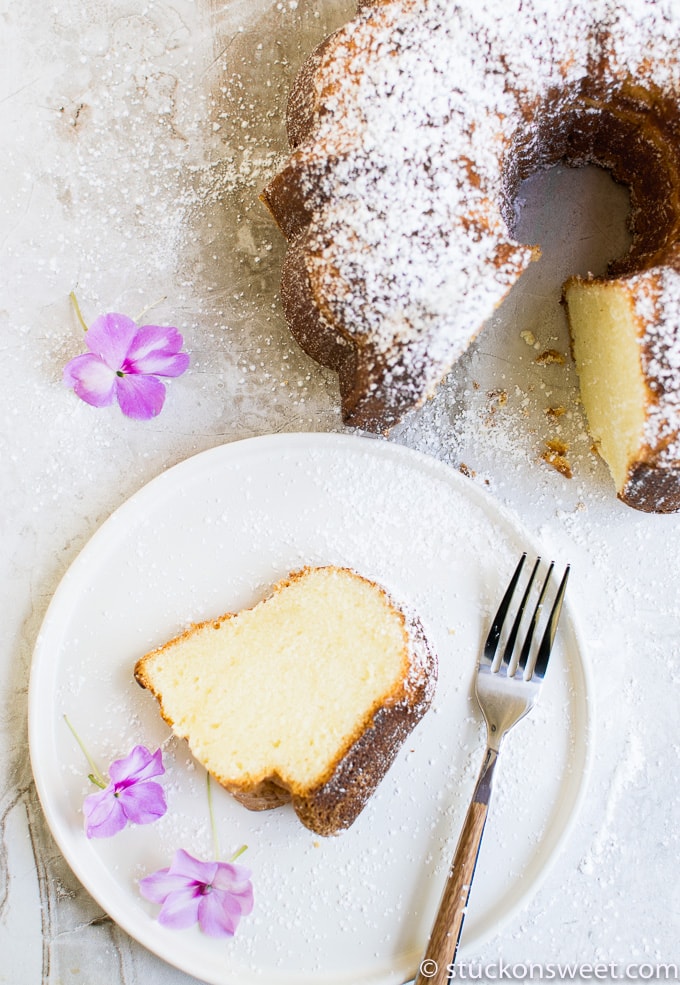 Soft vanilla pound cake dusted with powdered sugar, served on a white plate with pink flowers and a silver fork, showcasing homemade cake dessert from Stuck On Sweet.