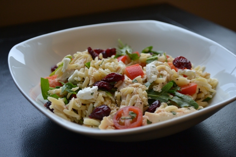 Fresh pasta salad with cherry tomatoes, arugula, feta cheese, dried cranberries, and a light dressing in a white bowl. Perfect for a healthy, flavorful meal or side dish.