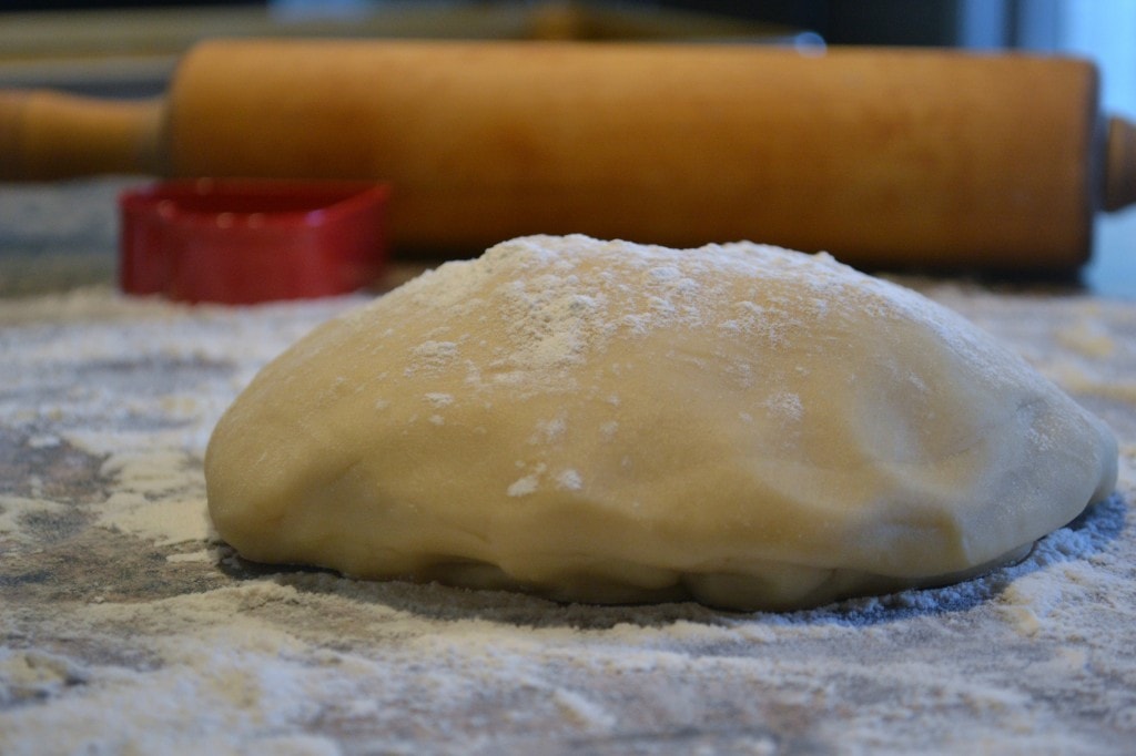 Soft sugar cookie dough ball dusted with flour, ready for baking or cooking, on a floured surface with a rolling pin and a measuring cup in the background.