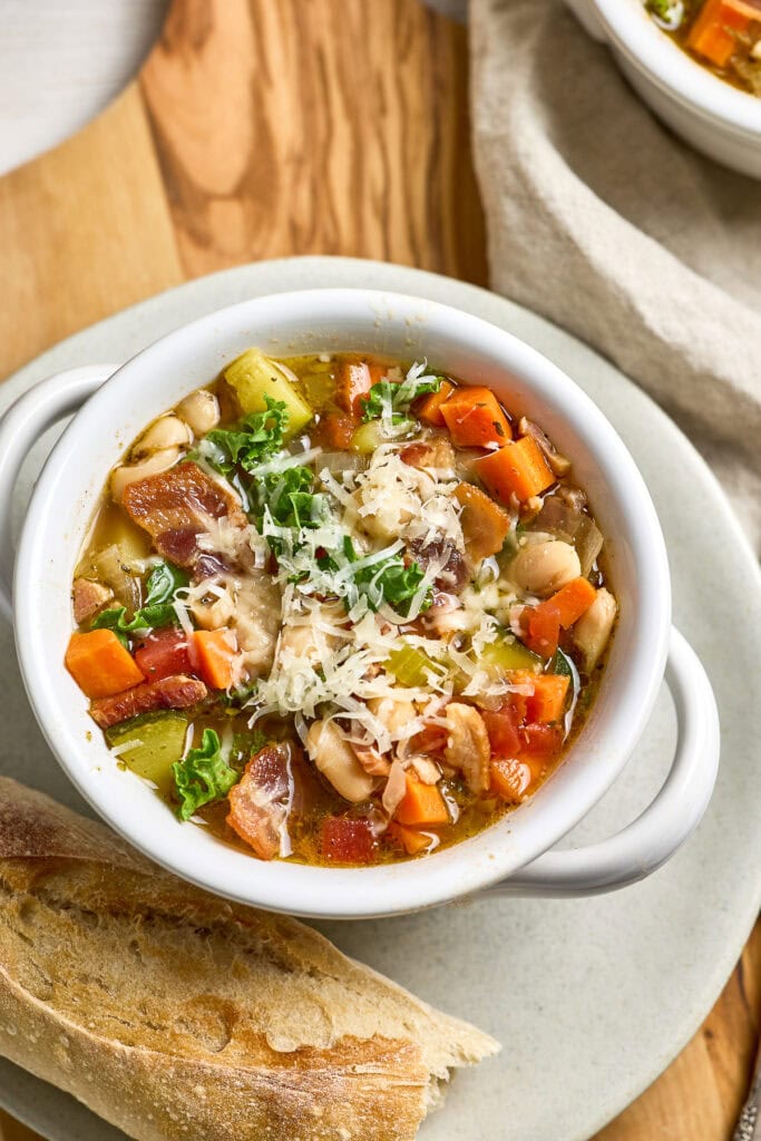 Tuscan Kale White Bean Soup in a white mug with shredded cheese, garnished with fresh herbs, served alongside a crusty baguette on a round stone plate.