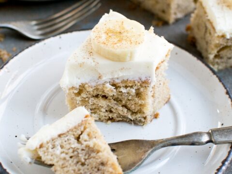 Creamy banana cake with white frosting topped with banana slices on a white plate. Soft, moist cake with smooth icing, perfect for desserts or special occasions.