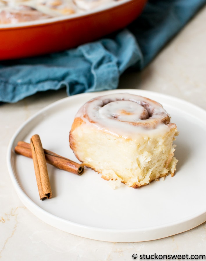 Rich cinnamon roll with white icing on a white plate, accented by cinnamon sticks, served as a sweet breakfast or dessert. Perfect for cinnamon roll lovers and bakery treats.