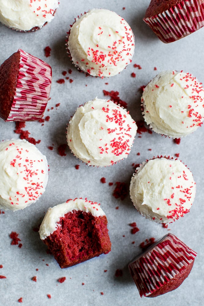 Red velvet cupcakes with cream cheese frosting and red sprinkles, topped on parchment paper, with some cupcakes in decorative wrappers, perfect for Valentine's Day or special occasions.