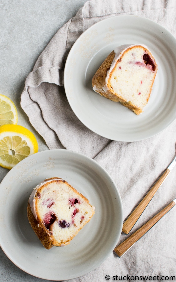 Lemon blueberry cake slice on white plate with lemon slices and wooden utensils, perfect for dessert or afternoon snack.