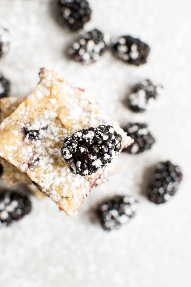 Blackberry lemon bar with powdered sugar on a marble surface, topped with a fresh blackberry. Delicious, fruity dessert perfect for summer or gift giving.
