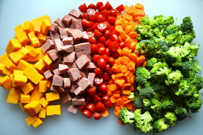 Fresh diced yellow cheese, cooked ham, cherry tomatoes, diced carrots, and broccoli florets, arranged in a colorful display for healthy meal prep or nutritious cooking.