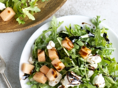 Fresh arugula salad with grilled chicken, crumbled cheese, balsamic glaze, and cantaloupe cubes on white plate. Perfect for a light, healthy meal.