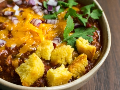 Creamy loaded chili with shredded cheese, chopped onions, cilantro, and crispy cornbread croutons in a bowl on a wooden table.