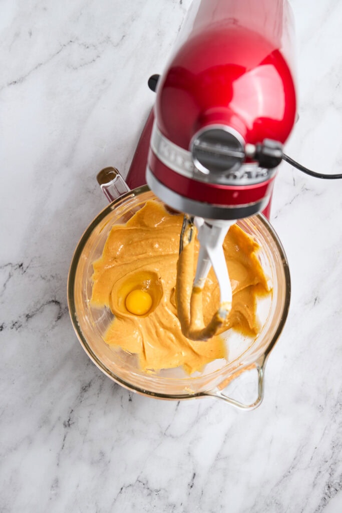 Creamy pumpkin cheesecake batter with an egg in a glass mixing bowl on marble countertop.