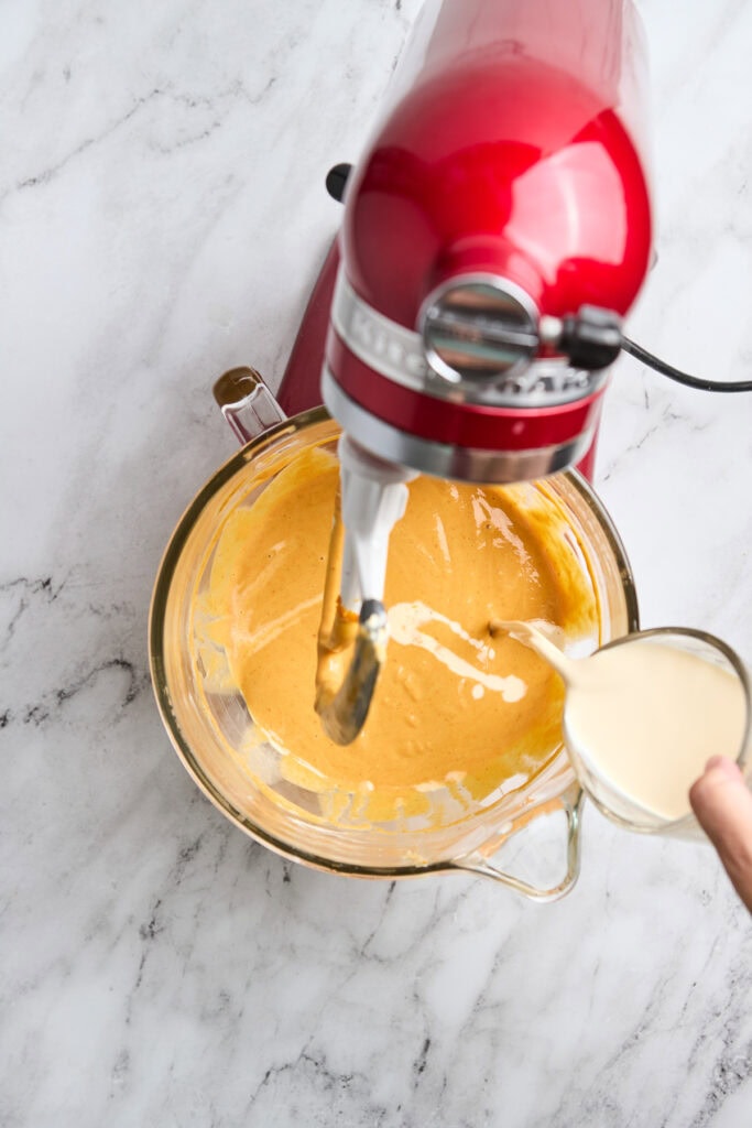 Cream-colored batter being mixed in a glass bowl with a red stand mixer on a marble surface.