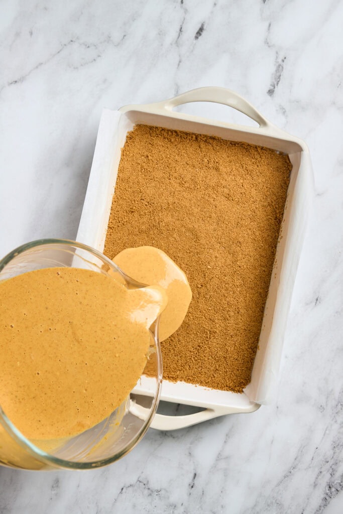 Creamy caramel sauce pouring into a glass bowl on a marble countertop with a graham cracker crust in a white baking dish.