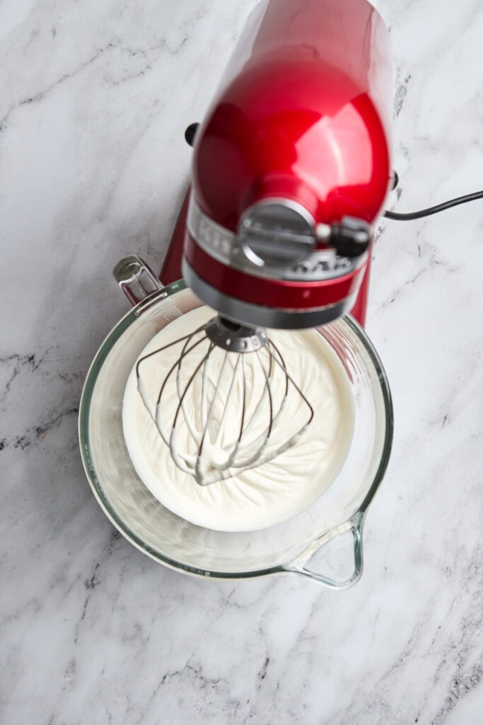 Fluffy whipped cream being mixed with a red stand mixer in a glass bowl for dessert or baking recipes.
