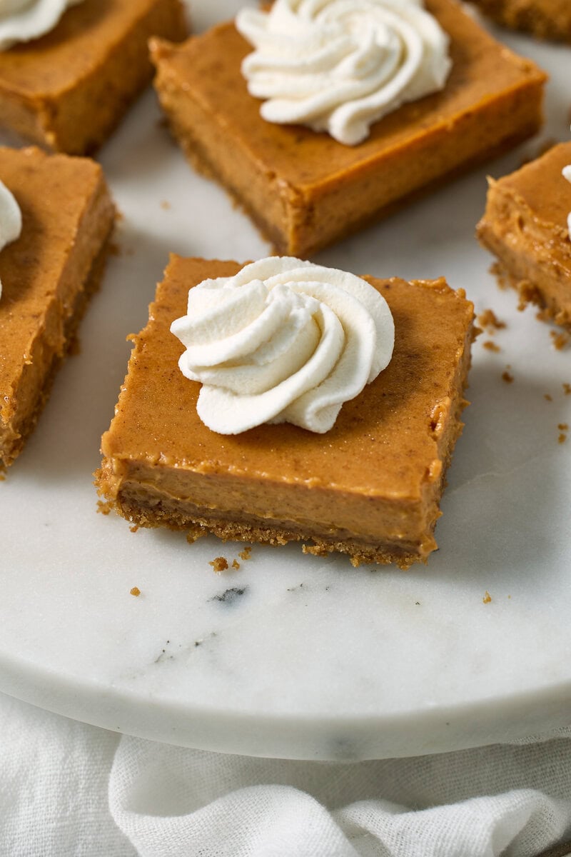 Cream cheese pumpkin bars topped with whipped cream on a white marble serving platter.