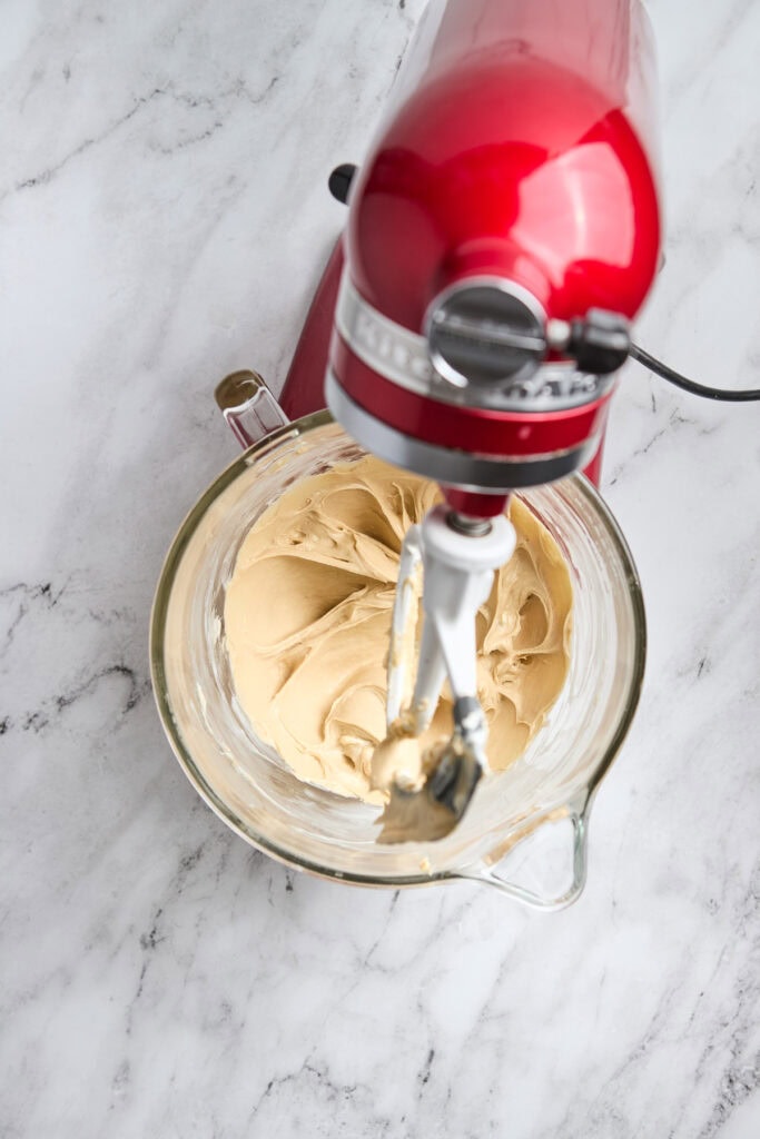Creamy vanilla frosting being mixed with a red stand mixer in a glass bowl.