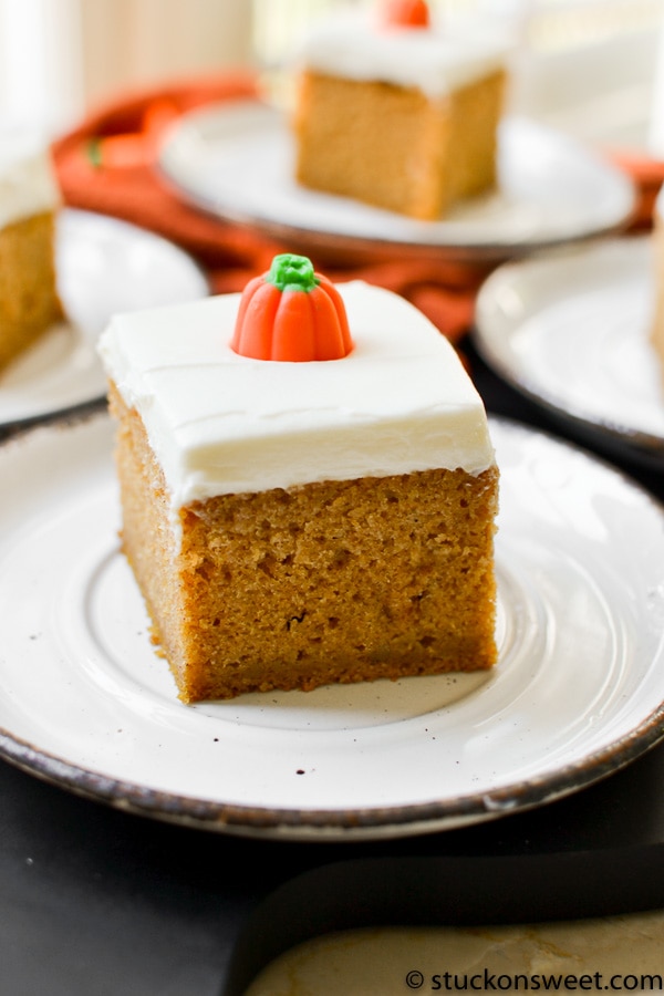 Bright pumpkin bars with cream cheese frosting and orange fondant decoration on top, served on a white plate. Perfect for holiday desserts and cake lovers looking for sweet, flavorful treats.