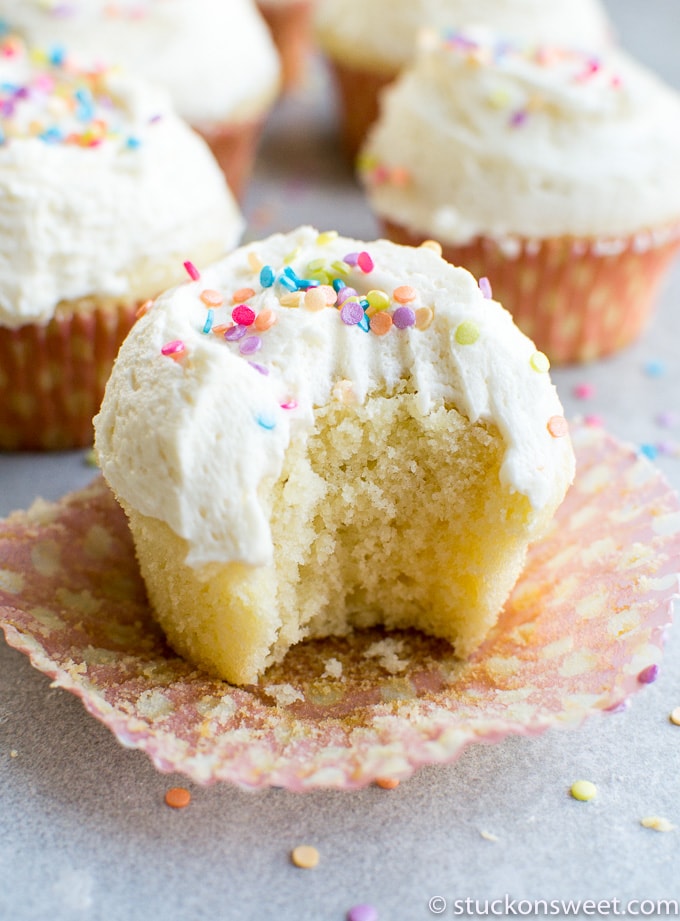 Soft vanilla cupcake with colorful sprinkles, partially eaten, on a parchment paper, with more cupcakes in the background; focus on the moist crumb and fluffy frosting.