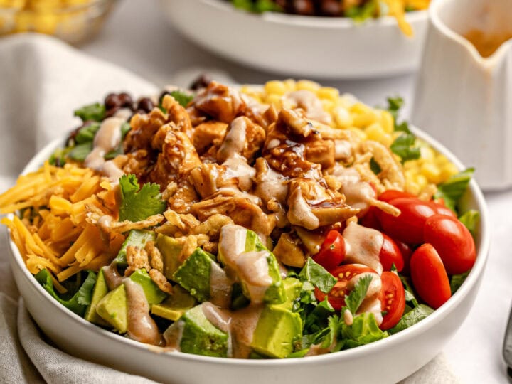 Fresh taco salad bowl with seasoned chicken, diced avocado, cherry tomatoes, shredded cheese, and creamy dressing on a white ceramic dish. Delicious and healthy meal option from Stuck On Sweet.