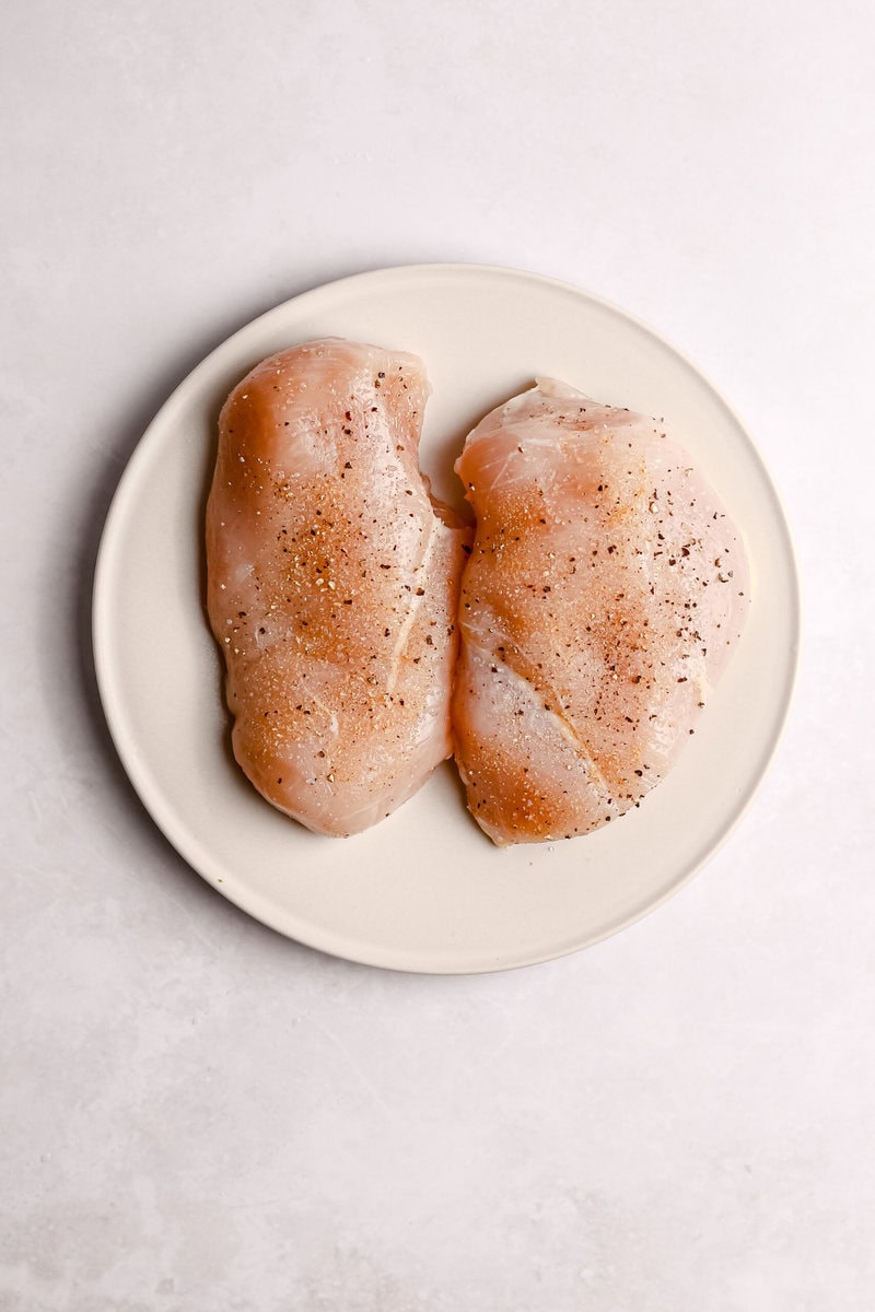 Raw chicken breasts seasoned with salt and pepper on a white plate.