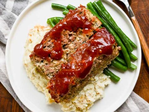 Homemade meatloaf with green beans and mashed potatoes on a white plate, served with ketchup. Classic comfort food recipe perfect for family dinners and easy weeknight meals.