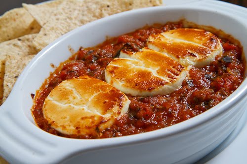Creamy baked brie cheese in a rich tomato and beef sauce served in a white ceramic dish with crispy bread slices on the side.