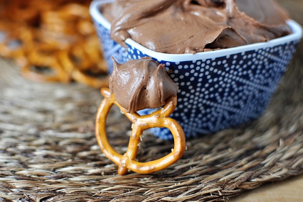 Creamy chocolate frosting in a blue patterned dish with pretzel and chocolate candies on a rustic woven surface.