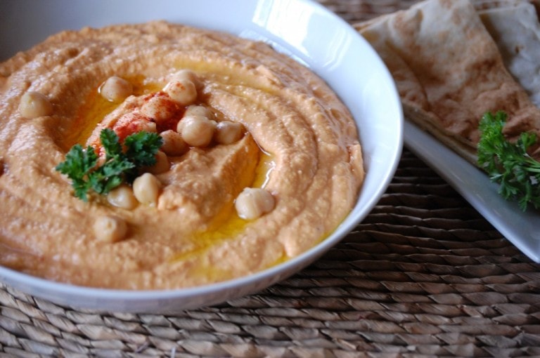 Creamy hummus with chickpeas and olive oil in a white bowl, garnished with parsley, served with flatbread on a woven placemat.