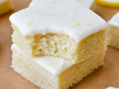 Soft lemon bars with white icing topping, fresh lemon slices in the background, on a brown parchment surface. Perfect for lemon dessert lovers, showcasing homemade citrus treats.