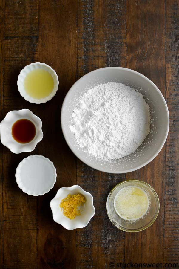 Finely ground powdered sugar, lemon zest, lemon juice, vanilla extract, maple syrup, and honey in small bowls with a large bowl of powdered sugar on rustic wooden table.