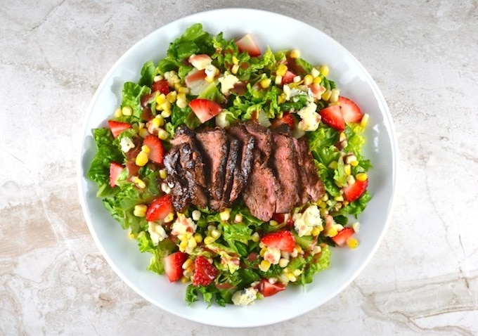 Grilled steak salad with fresh strawberries, corn, blue cheese, and greens, served on a white plate, healthy and flavorful summer dish from Stuck on Sweet.