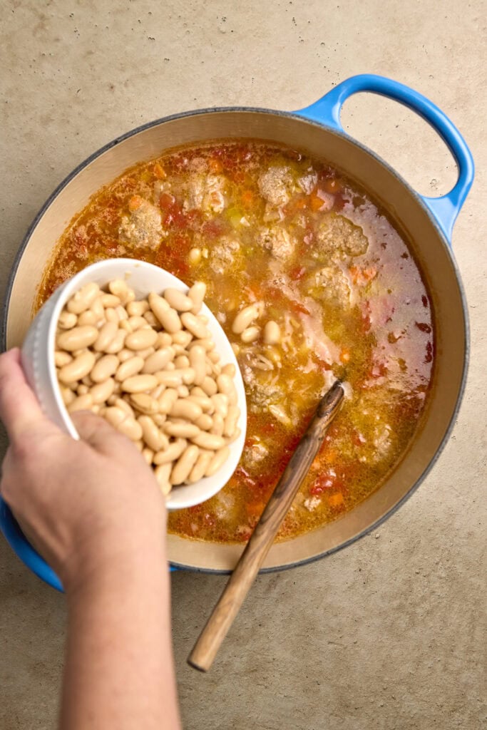 Creamy white beans being added to a flavorful homemade soup in a large pot.