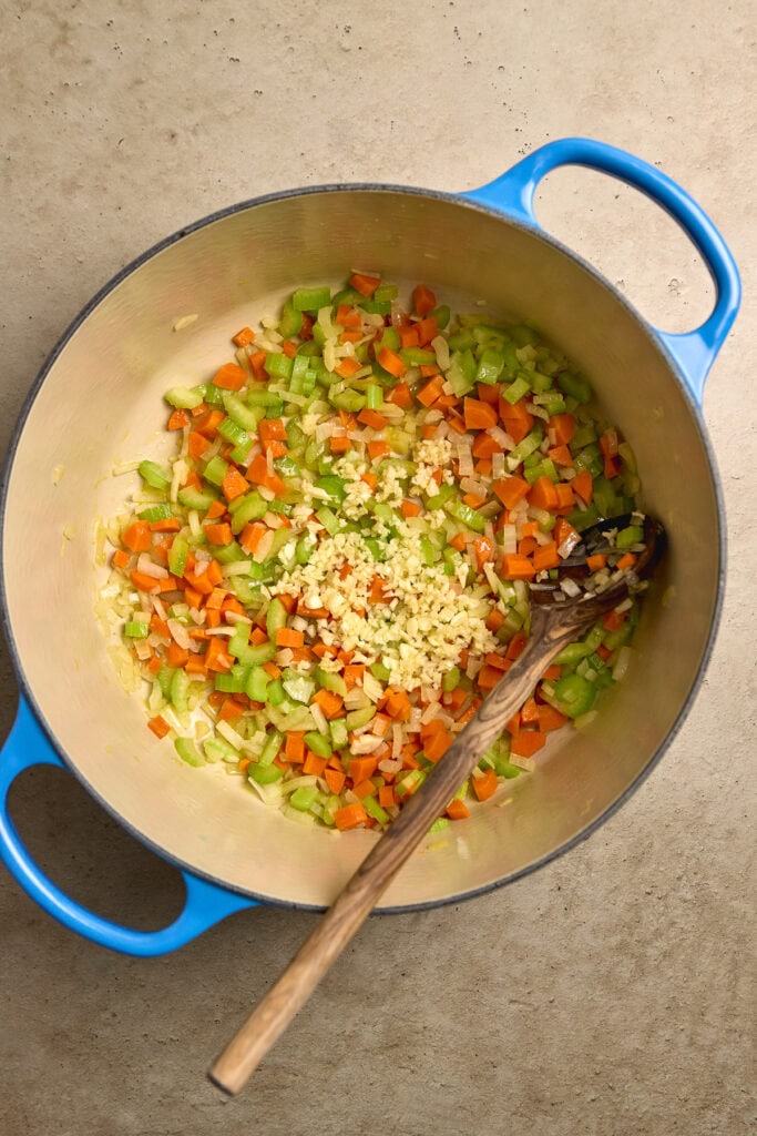 Diced celery, carrots, and garlic being sautéed in a small stainless steel pot with blue handles for a homemade vegetable soup or stew.