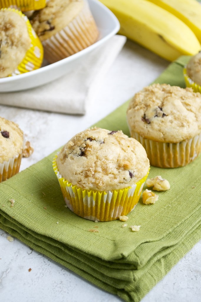 Moist banana chocolate chip muffins on a green napkin with a bowl of muffins and bananas in the background.