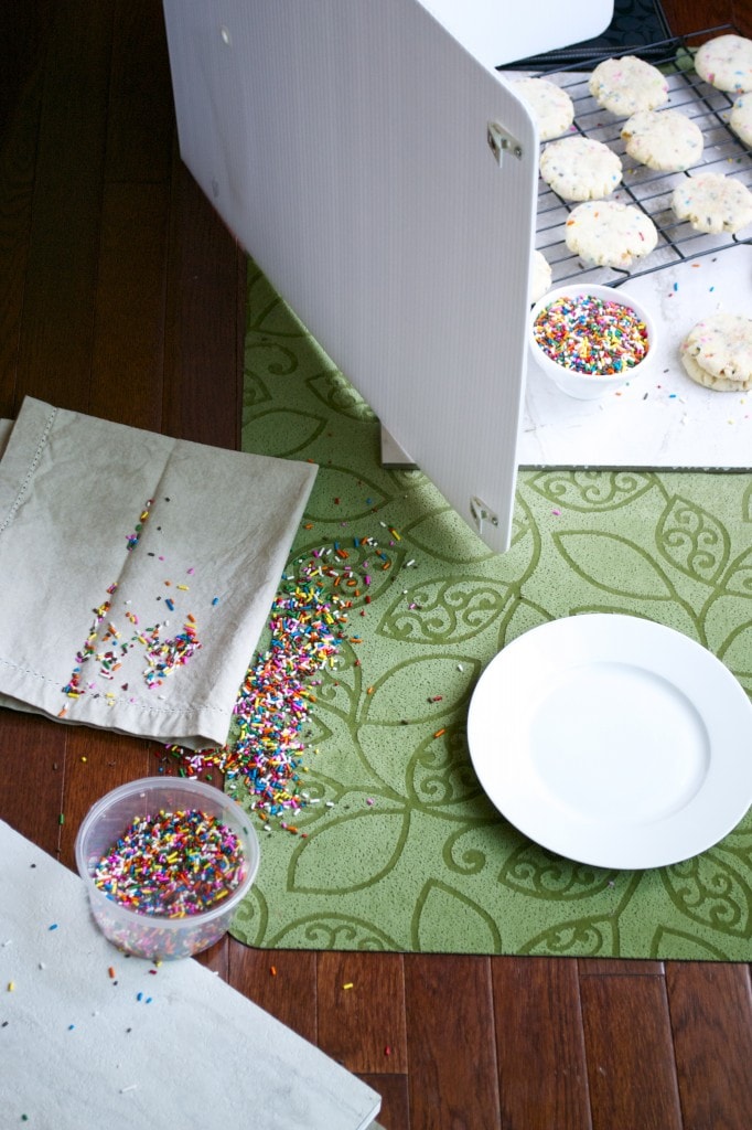 Colorful sprinkles scattered on napkin and table, preparing for baking cookies, with cookies cooling on a rack and a white plate nearby.