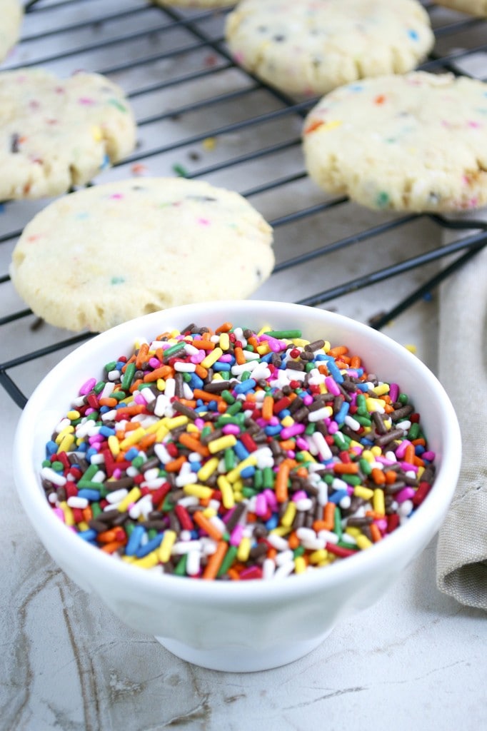 Colorful rainbow sprinkles in a white bowl with sugar cookies with rainbow sprinkles on a cooling rack in the background.