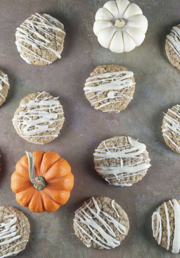Pumpkin-shaped cookies with white chocolate drizzle and small decorative pumpkins on a rustic surface, perfect for fall desserts and Halloween treats.