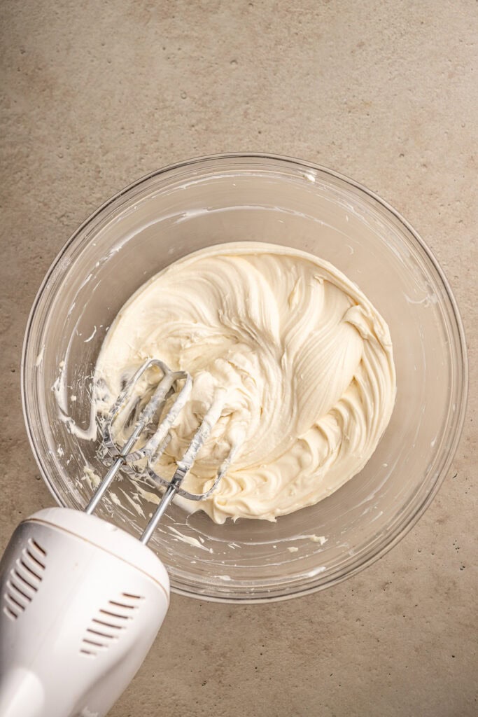Cream cheese frosting being mixed in a clear glass bowl with an electric hand mixer on a beige surface.