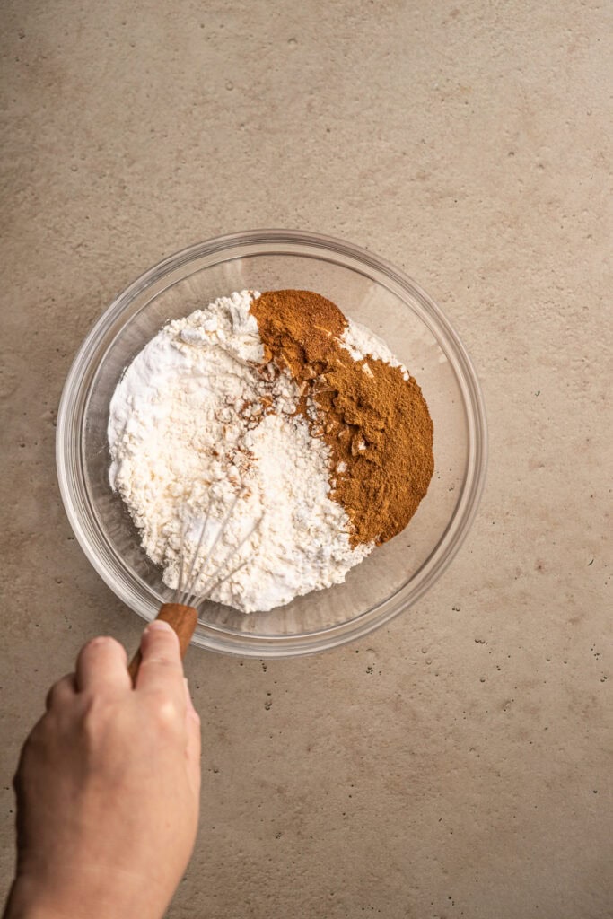 Cream-colored flour and brown cinnamon in a glass mixing bowl, with a hand whisking the ingredients, on a neutral surface.