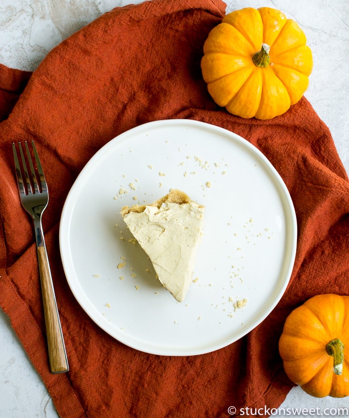 Creamy pumpkin cheesecake slice on a white plate surrounded by mini pumpkins, with a rustic orange cloth and fork for a festive fall dessert presentation.