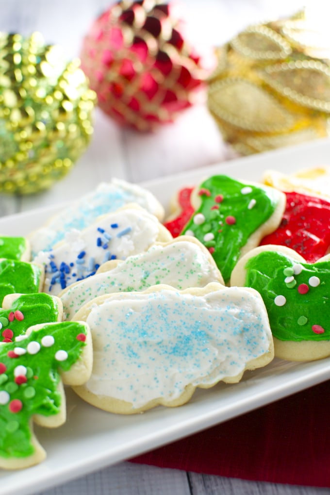 Festive Christmas sugar cookies decorated with colorful icing and sprinkles on a white platter with holiday ornaments in the background.