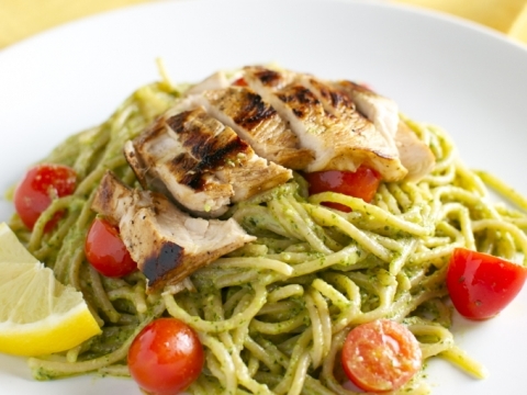 Sliced grilled chicken atop creamy pesto pasta with cherry tomatoes and lemon wedges on white plate, close-up, fresh and delicious Italian-inspired dish.