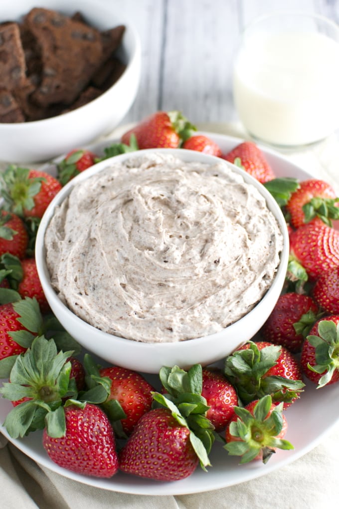 Creamy chocolate chip cookie dough dip served in a white bowl surrounded by fresh strawberries and a glass of milk on a rustic wooden table. Perfect for snacks or parties.