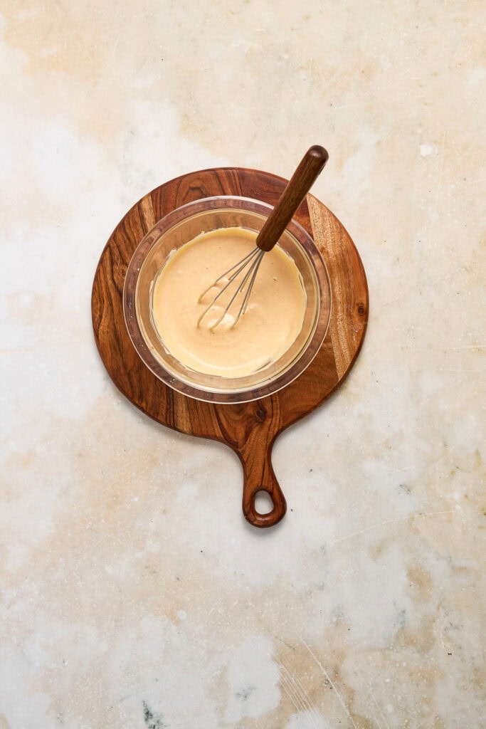 Creamy cheese sauce in a glass bowl with a whisk, served on a wooden board, perfect for cheeseburger quesadillas.