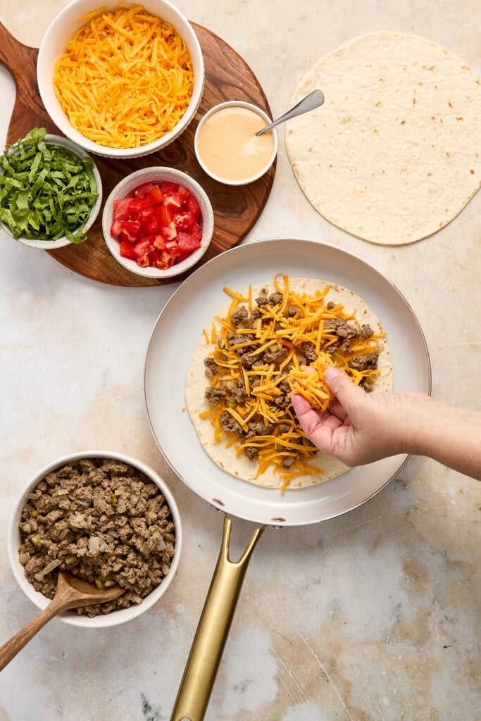 Delicious cheeseburger quesadilla being assembled with shredded cheese, seasoned ground beef, and fresh toppings like tomatoes and lettuce on a white plate.