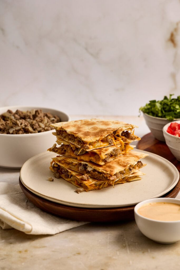 Close-up of cheesy cheeseburger quesadillas stacked on a plate, with sides of fresh salad and dipping sauce, perfect for a savory snack or meal.