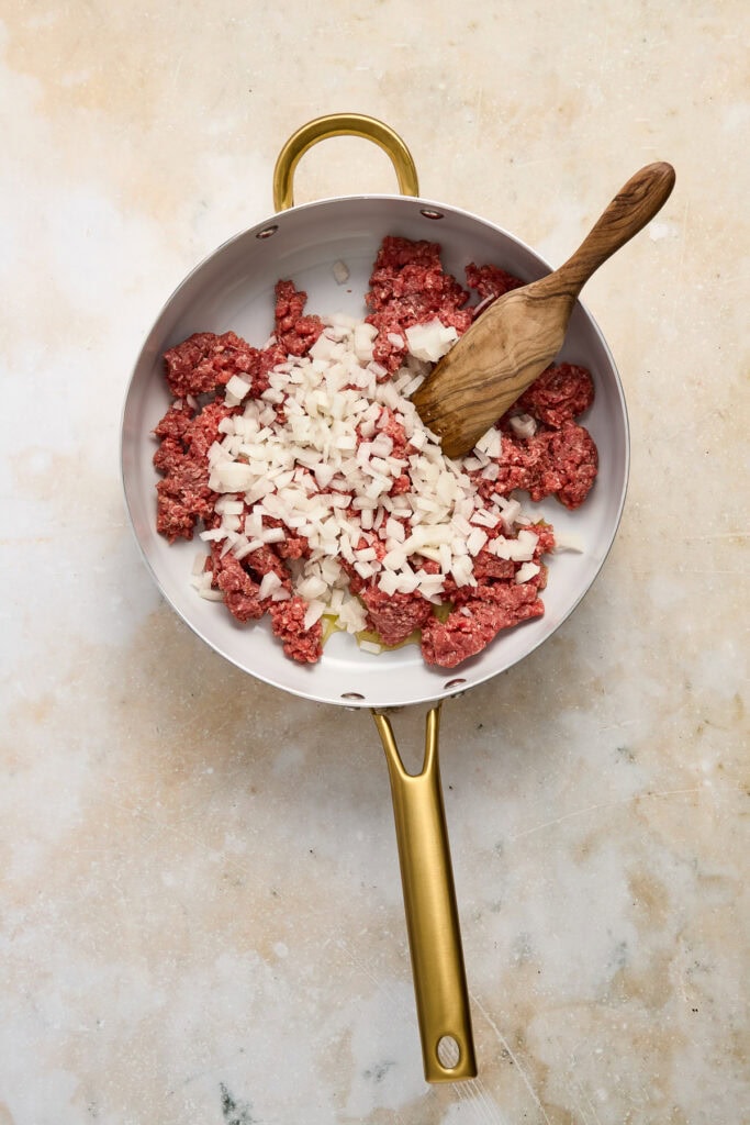 Ground beef and chopped onions in a skillet, ready to make cheeseburger quesadillas, with a wooden spoon for stirring.