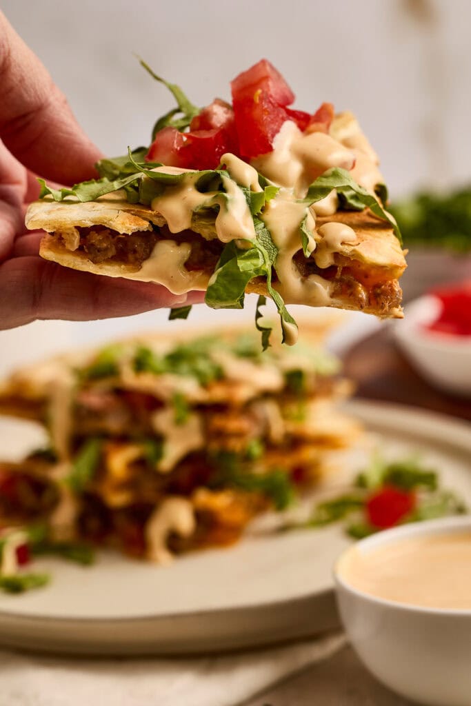 Close-up of a cheeseburger quesadilla topped with fresh tomatoes, lettuce, and creamy sauce, held above a plate with more quesadillas and dipping sauce.