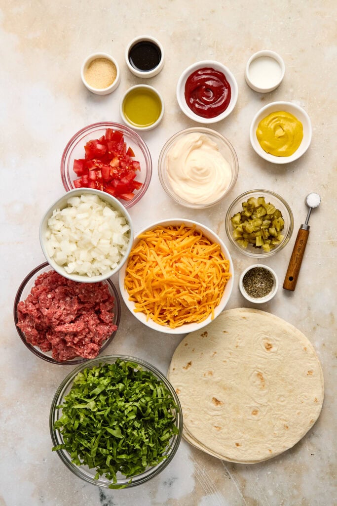 An image showcasing ingredients for cheeseburger quesadillas, including ground beef, shredded cheese, diced onions, chopped pickles, tomato, mayonnaise, ketchup, mustard, and flour tortillas, arranged.