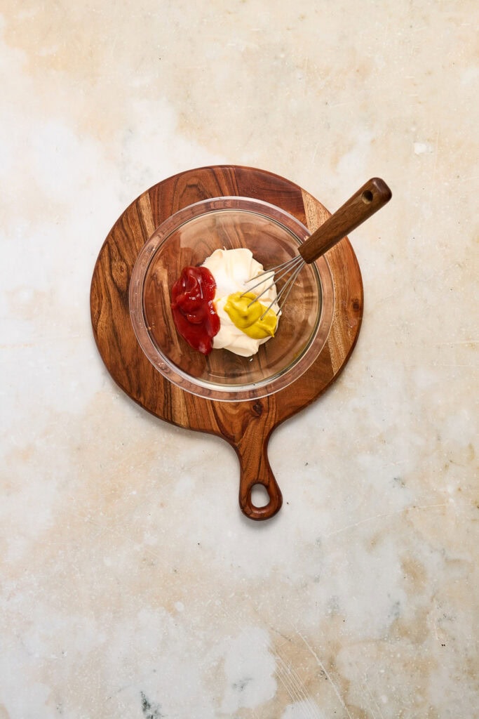 Creamy mayonnaise, ketchup, and mustard in a glass bowl with a whisk on a wooden cutting board, ready for cheeseburger quesadillas.