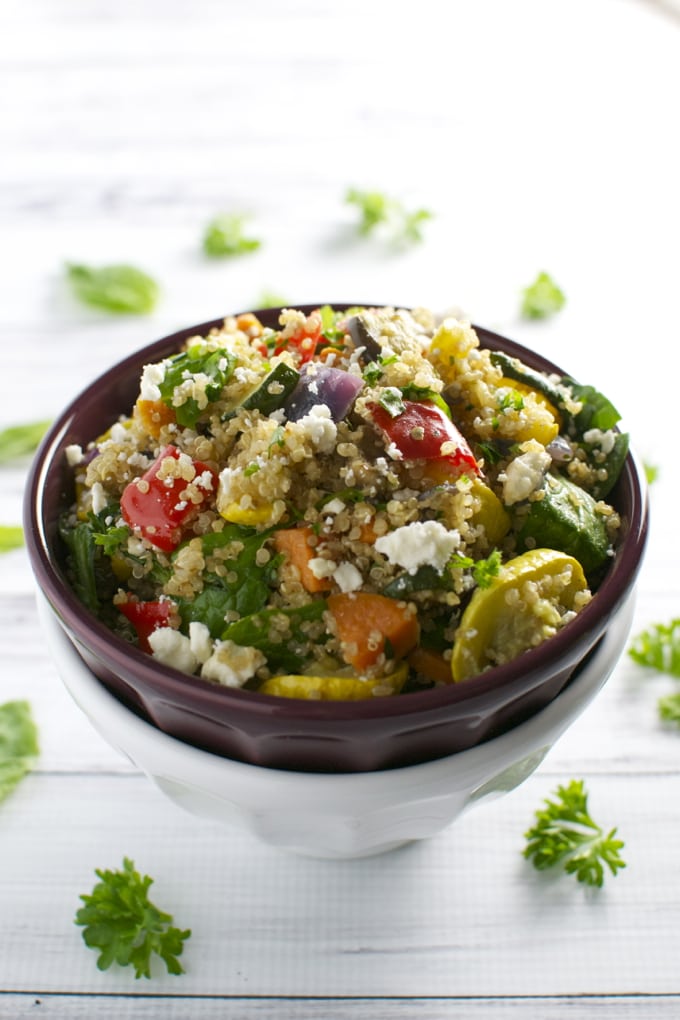 Colorful quinoa salad with fresh vegetables and crumbled feta cheese in a dark bowl on white wooden background.